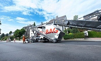 Foto einer Asphaltbaumaschine mit der Aufschrift STRABAG, sie steht am Straßenrand, der Himmel ist bewölkt