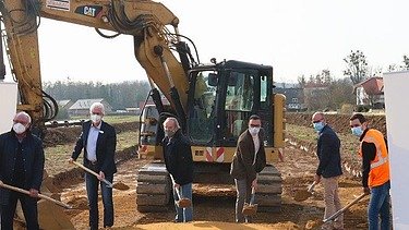 Foto von mehreren Personen beim symbolischen Spatenstich mit Schaufeln in der Hand, dahinter ein großer, gelber Bagger