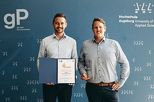 Foto von zwei Personen vor blauem Hintergrund, die linke Person hält eine Urkunde