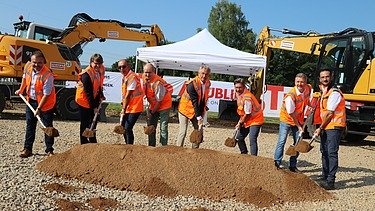 Foto von mehreren Personen bei sonnigem Wetter auf einer Baustelle, die jeweils einen Spaten in den Händen halten und einen symbolischen ‚Spatenstich‘ ausführen