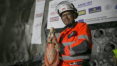Foto eines Tunnelbauarbeiters in Schutzkleidung, er hält eine Madonna Figur in den Händen