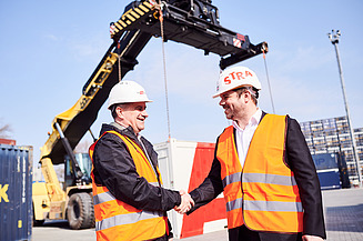 Foto von zwei Personen, die sich die Hand schütteln und Schutzkleidung tragen, dahinter sieht man einen Bagger mit ausgefahrenen Greifarm