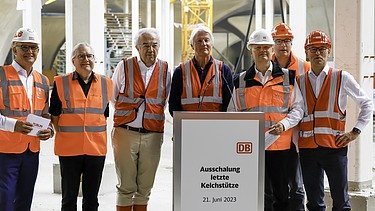 Foto von mehreren Personen in Schutzausrüstung die vor einem Rednerpult stehen. 
