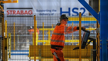 Foto eines Arbeiters hinter einem Bauzaun, der eine Maschine bedient