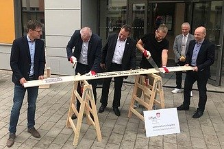 Foto von mehreren Männern im Anzug, die ein Brett sägen. 