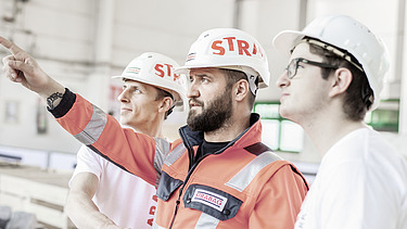 Das Bild zeigt drei Personen mit weißen Bauhelmen mit der Aufschrift STRABAG