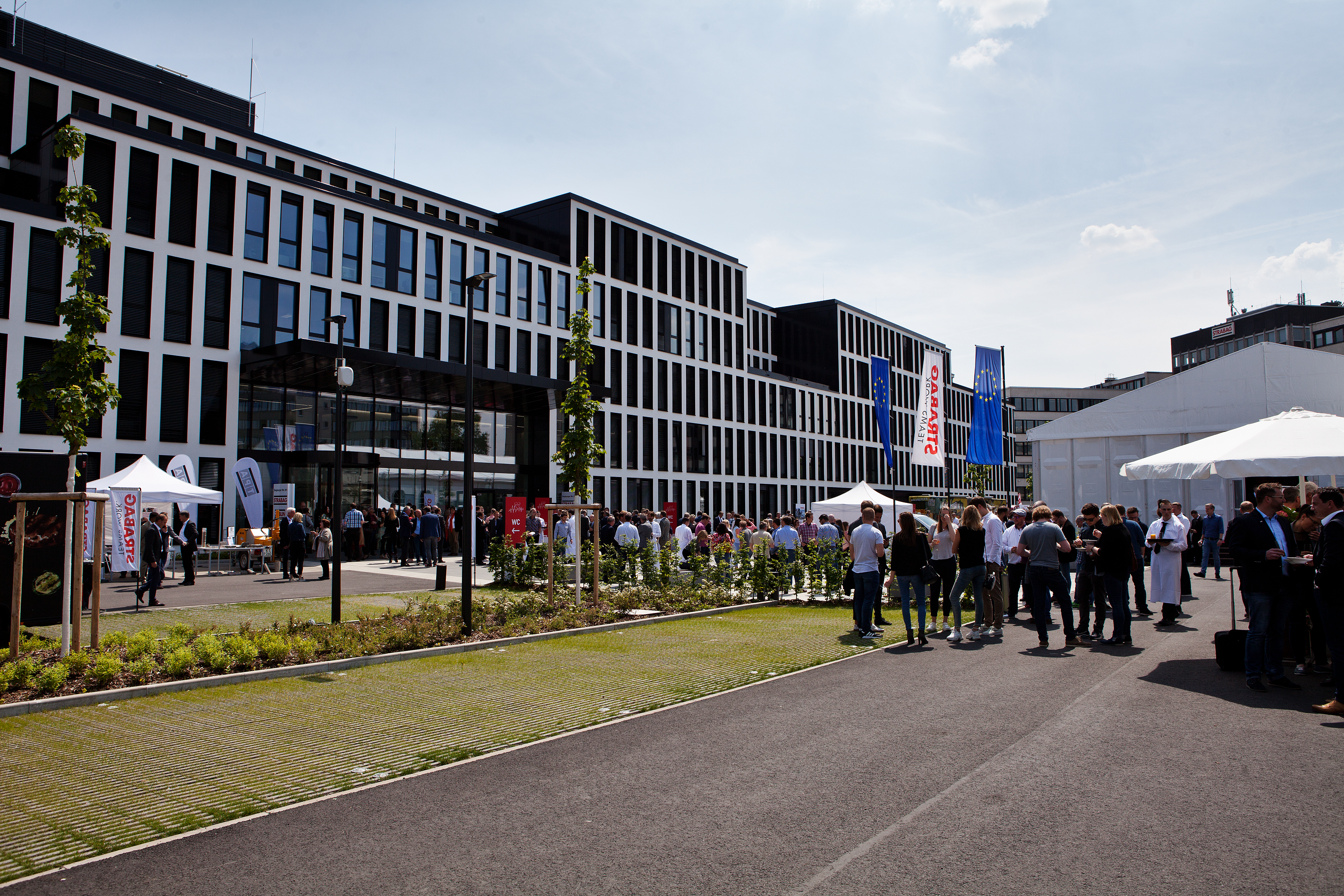 Foto vom Unternehmenszentrale in Köln auf dem Parkplatz. Es ist ein Zelt aufgebaut und Menschen feiern die neue Zentrale.   