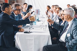 Foto von mehreren Personen bei der Jubiläumsfeier am Standort Sande. Sie sitzen an einem Tisch und stoßen an. 