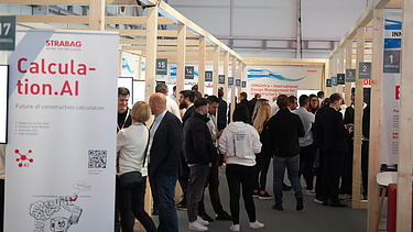 Photo of several people between exhibition stands, in the foreground on the left is a roll-up