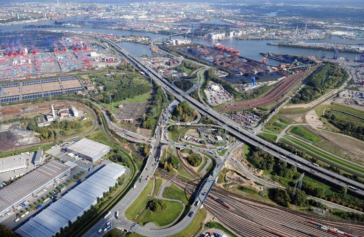 Luftaufnahme von einem Autobahn-Drehkreuz, links steht eine große Lagerhalle, im Hintergrund ist ein Hafen zu sehen