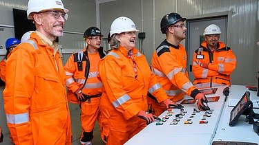 Photo of several people wearing hard hats and orange protective clothing standing at a control panel