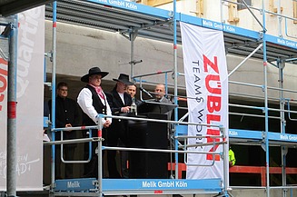 Rede zum Richtfest auf einem Gerüst auf der Baustelle im Antoniter Quartier in Köln, gehalten vom Polier. Um ihm herum sind Männer. 