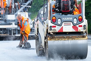 Foto einer Straßenwalze, dahinter steht ein Arbeiter mit einer Schaufel