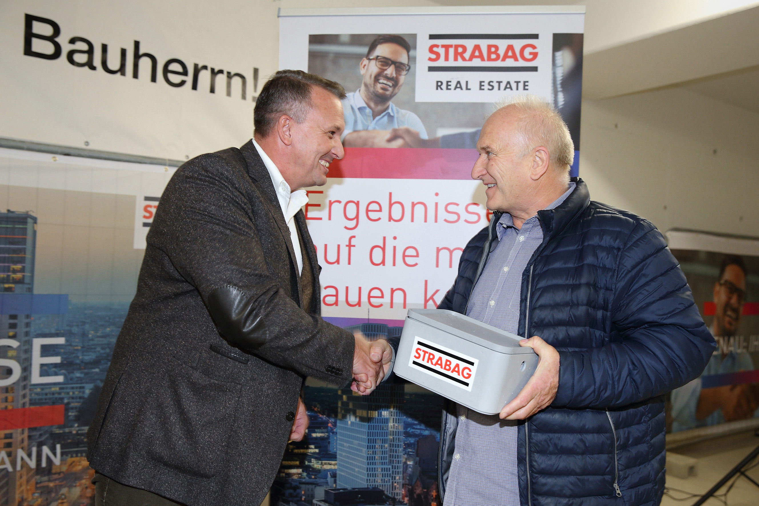 Foto von zwei Personen, die sich die rechte Hand schüttel, die Person rechts hält eine Geldschatulle in der linken hand