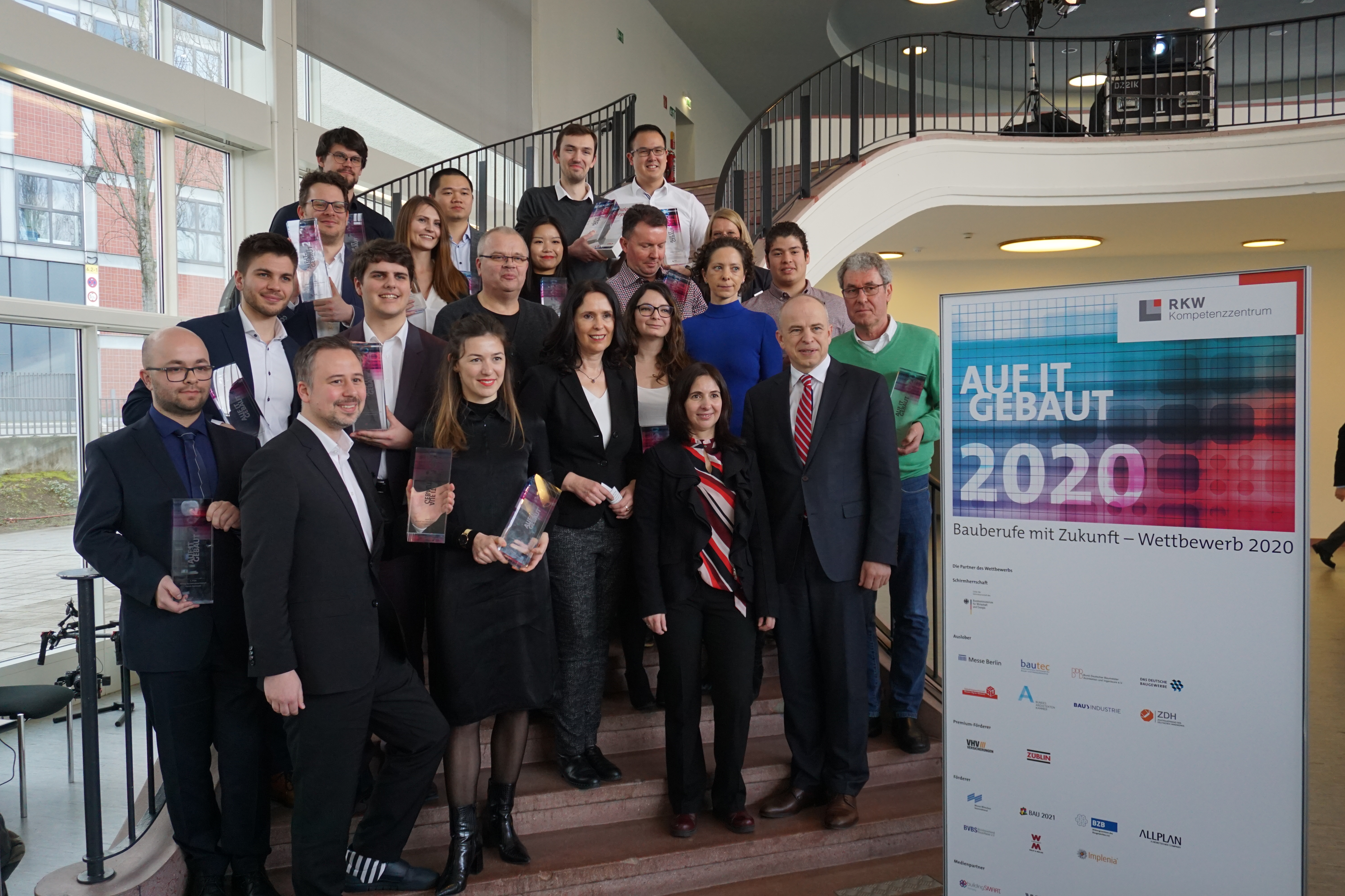 Foto von mehreren Personen auf einer Treppe. Sie sind schick angezogen und halten ein Pokal in den Händen. Neben ihnen ist ein Banner von Bauberufe mit Zukunft – Wettbewerb 2020.     