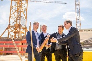 Foto von vier Männern in Anzug auf einer Baustelle. Sie halten eine Röhre. 