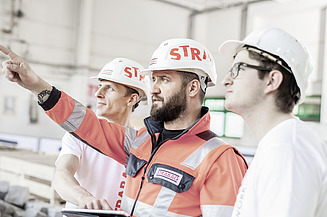 Das Bild zeigt drei Personen mit weißen Bauhelmen mit der Aufschrift STRABAG
