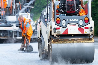 Foto einer Straßenwalze, dahinter steht ein Arbeiter mit einer Schaufel