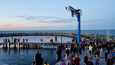 Eine Hand voll Menschen befindet sich auf einer Seebrücke. Die Seebrücke ist abgerundet und ein blaue Säule steht in der Mitte, sie ebnet den Weg zu einem Steg, der in die Mitte der Seebrücke führt.