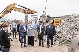 Foto von mehreren Personen bei einer Baustellenbesichtigung, im Hintergrund ein gelber Bagger bei der Arbeit