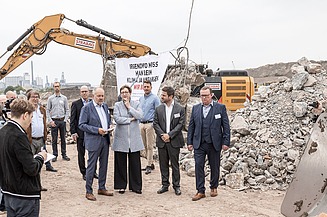 Foto von mehreren Personen bei einer Baustellenbesichtigung, im Hintergrund ein gelber Bagger bei der Arbeit