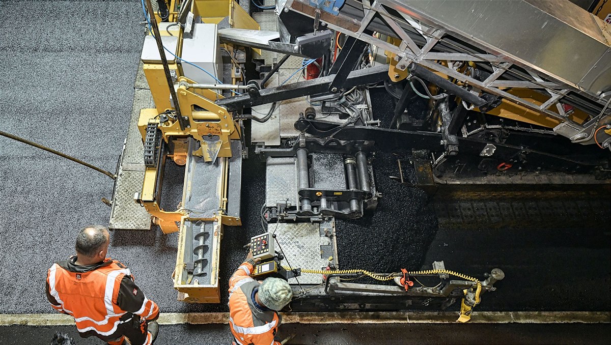 Foto zeigt zwei Straßenbauarbeiter in Schutzwesten vor einer Asphaltbaumaschine