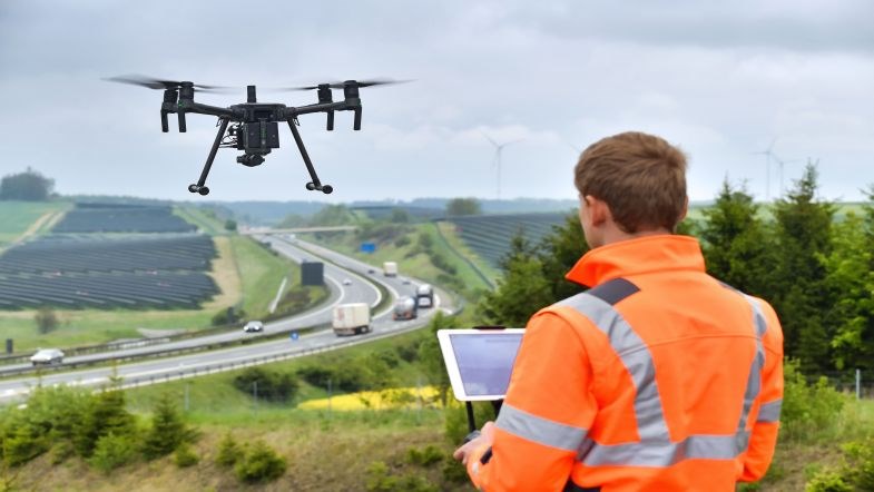 Foto einer Person in Schutzjacke mit einem Tablet in der Hand bedient er eine fliegende Drohne